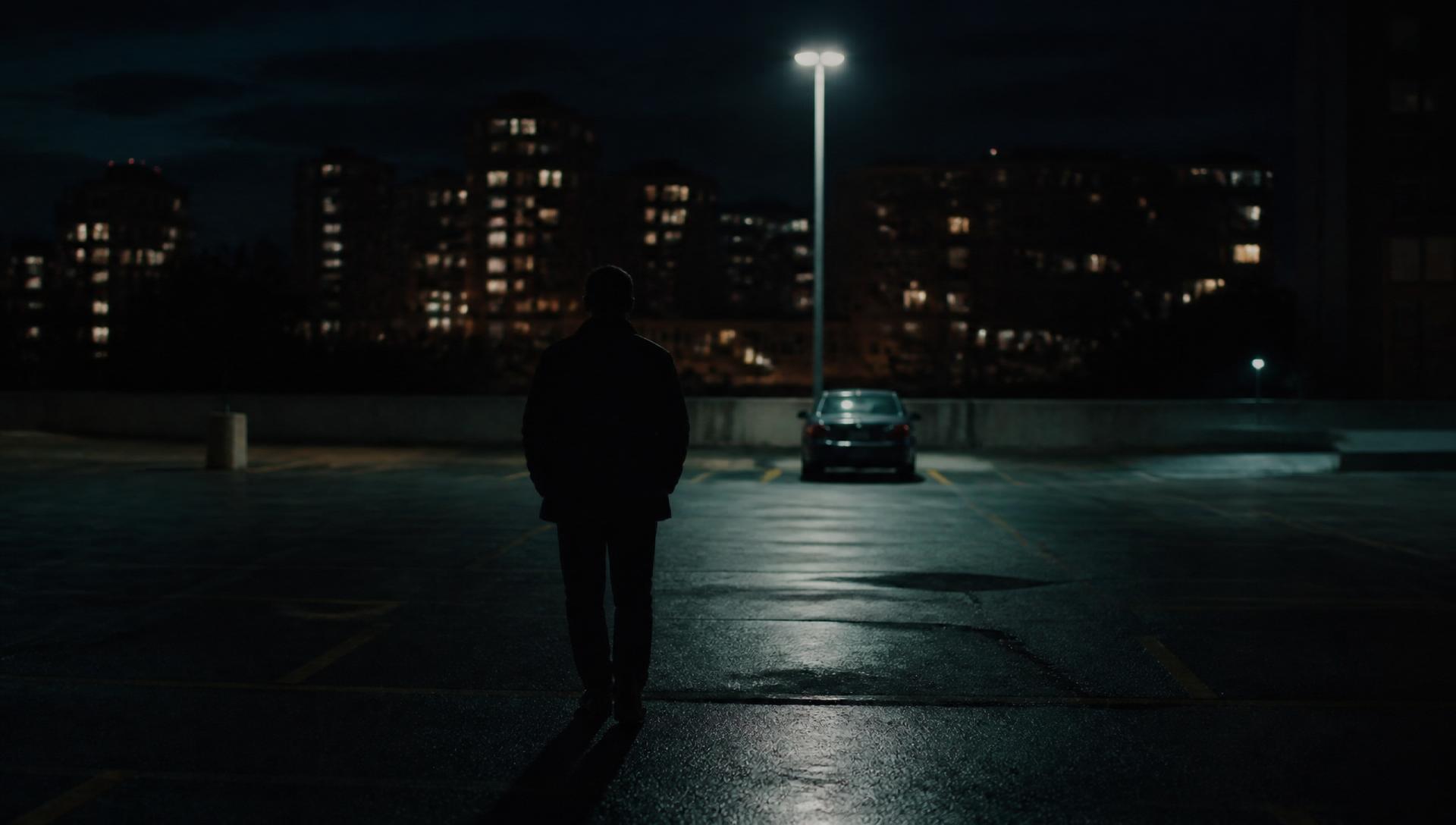 A lone figure walking across a dim parking lot at night toward a distant car.